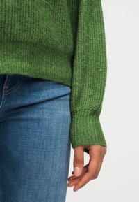 Personne portant un pull vert en tricot avec des poignets côtelés et un jean en denim bleu, main détendue sur le côté contre un fond uni.