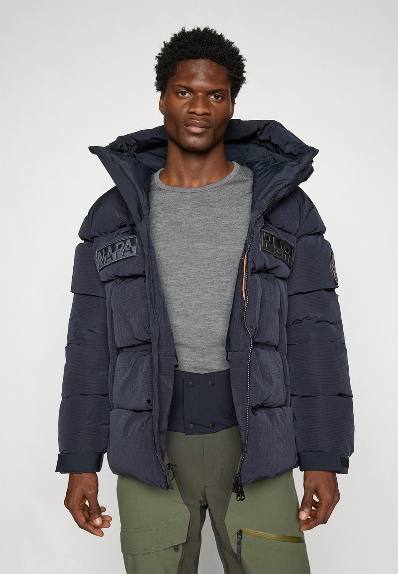 Veste matelassée marine avec capuche, présentant des sections en losange, des écussons avec logo sur la poitrine et une fermeture éclair. Portée par-dessus un T-shirt gris.