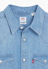 Chemise en denim bleu clair avec col pointu, boutons-pression, poches poitrine et étiquettes visibles de la marque Levi's.