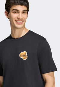 Černé bavlněné tričko s malou grafikou tří zlatých mincí na prsou. Krátké rukávy a kulatý výstřih.