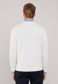 Maglione bianco a maglia con maniche lunghe e orlo a costine, indossato sopra una camicia azzurra con colletto, con un dettaglio del logo sull'orlo.