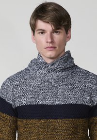 Gehaakte trui met een hoge kraag, met horizontale strepen in grijs, marineblauw en mosterdkleuren, die verschillende texturen en patronen tonen.