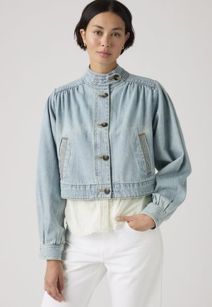 Frau trägt hellblaue Jeansjacke mit Knöpfen und hohem Kragen über weißer Spitzenbluse und weißer Hose, steht vor einem einfarbigen Hintergrund.