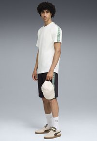 Junger Mann, der ein weißes T-Shirt und schwarze Shorts mit grünen Mustern an den Seiten trägt, hält eine beige Mütze in der Hand und steht vor einem grauen Hintergrund.