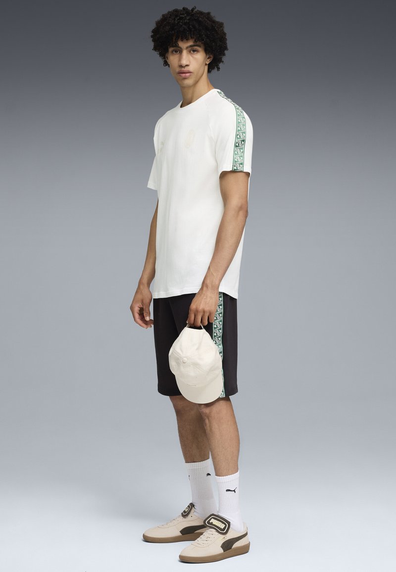 Junger Mann, der ein weißes T-Shirt und schwarze Shorts mit grünen Mustern an den Seiten trägt, hält eine beige Mütze in der Hand und steht vor einem grauen Hintergrund.