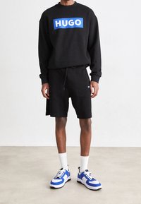 Felpa nera con logo "HUGO" blu, abbinata a pantaloni corti neri. Presenta calze bianche alla caviglia e sneakers blu e bianche con dettagli neri.