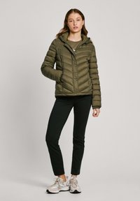 Olijfgroene pufferjack met een capuchon, voorzien van diagonale stiksels, drukknoopsluitingen en zijzakken, gedragen met zwarte broeken en sneakers.