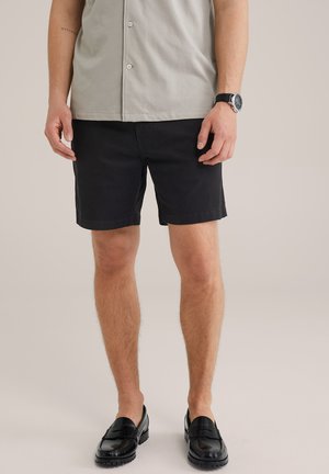 Shorts noirs en tissu lisse, avec une coupe droite et des poches latérales, portés avec une chemise bouton-Down gris clair et des mocassins noirs.