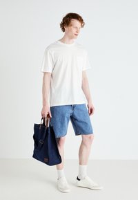 Witte katoenen t-shirt met zak, blauwe denim shorts en een donkerblauwe totebag met bruine leren accenten. Witte sokken en sneakers.