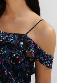 Blouse fleurie avec design à épaules dénudées, présentant un fond bleu marine et des motifs vibrants en violet, turquoise et blanc. Tissu léger avec détail volanté.