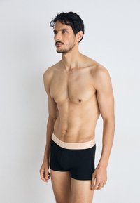 Boxer shorts noirs avec une taille beige clair arborant un subtil logo Lacoste. Le tissu semble lisse et ajusté.