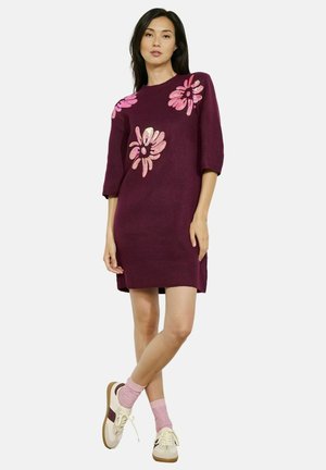 Abito lavorato a maglia bordeaux con maniche corte, caratterizzato da applicazioni floreali rosa. Abbinato a calzini rosa e sneaker beige, evidenziando un look casual.