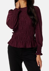 Mujer vistiendo una blusa burdeos de manga larga con fruncido en el dobladillo y pantalones negros, con la mano apoyada en el cuello.