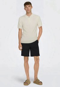 Polo shirt beige clair à manches courtes avec un col zippé, associé à un short noir. Le modèle porte des sandales marron. Design minimaliste avec une texture lisse.