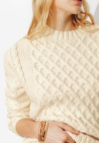 Pull en tricot couleur crème avec un motif en losanges texturé et un col côtelé. Des bracelets dorés au poignet ajoutent des accents métalliques.