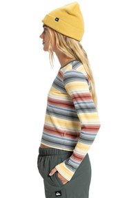 Gestreepte lange mouwen top met gele, rode, grijze en blauwe lijnen, gecombineerd met losse groene broeken. Een gebreide gele beanie maakt de look compleet.