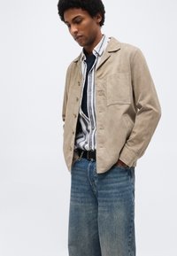 Homme portant une veste beige boutonnée par-dessus une chemise rayée et une ceinture noire avec un jean bleu, debout les mains dans les poches, regardant vers le bas.