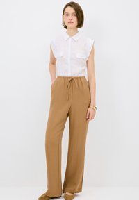 Witte mouwloze button-up shirt met borstzakken, gecombineerd met beige gestructureerde wijde broek. Bruine instapschoenen, effen achtergrond.
