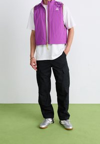 Fioletowa kurtka bez rękawów z zamkiem błyskawicznym, dwoma bocznymi kieszeniami i regulowanymi sznurkami przy ramionach, zestawiona z czarnymi spodniami typu cargo.