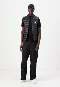 Melnā pūkainā veste ar rāvējslēdzēju priekšā, kombinēta ar melnu T-kreklu un kravas biksēm. Balti sporta apavi noslēdz apģērbu. Modelis stāv pretī baltai fona.