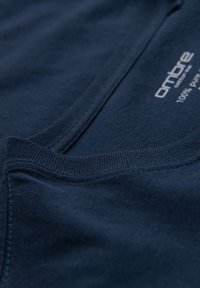 Nærbillede af marineblåt stof med synlig søm og ribbet halsudskæring, der viser en hvid tekstlabel med "ombre" og "100% ren bomuld."