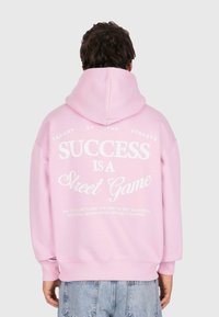 Persona che indossa un cappuccio rosa con testo bianco sulla schiena: "IL SUCCESSO È UN GIOCO DI STRADA", abbinato a jeans azzurri chiari.