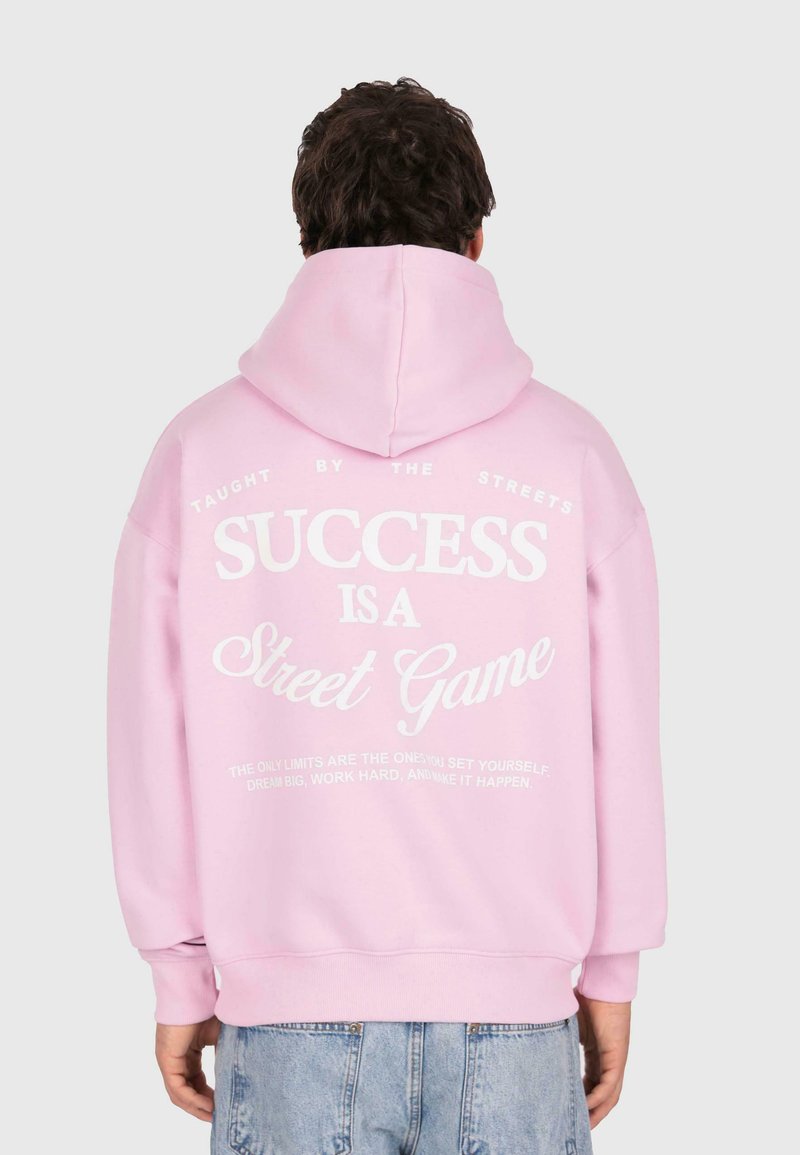 Persona che indossa un cappuccio rosa con testo bianco sulla schiena: "IL SUCCESSO È UN GIOCO DI STRADA", abbinato a jeans azzurri chiari.