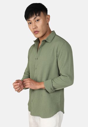 Grünes, strukturiertes Langarmhemd mit Button-Down-Kragen, das ein dezentes Muster aufweist und mit hochgekrempelten Ärmeln getragen wird, kombiniert mit weißen Hosen.