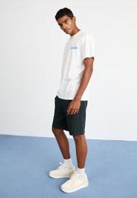 White cotton t-shirt, short sleeves, featuring a turquoise logo on the chest. Paired with dark green shorts and light-colored sneakers.