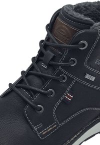 Botte en cuir noir avec doublure en laine, œillets en métal et couture blanche contrastante. Présente une patch en cuir brun et des accents texturés.