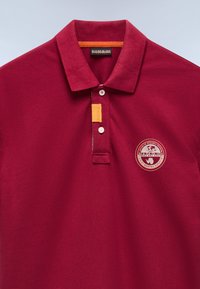Polo shirt bordeaux con colletto, tre bottoni bianchi e una toppa arancione. Presenta un logo ricamato circolare sulla sinistra.