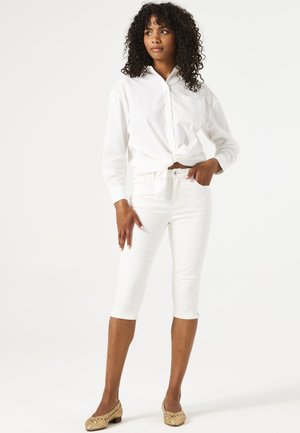 Chemise boutonnée en coton blanc nouée à la taille, associée à un jean blanc court. Le mannequin se tient en sandales tressées beiges devant un fond uni.