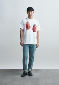 Λευκό βαμβακερό t-shirt με γραφικά αποτυπώματα κόκκινων γαντιών πυγμαχίας. Φορεμένο με ανοιχτό μπλε τζιν και μαύρα μοκασίνια. Απλός σχεδιασμός και άνετη εφαρμογή.