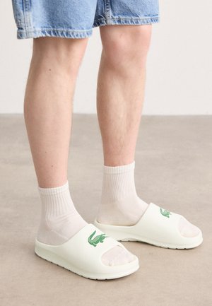 White slide sandals with an open toe, featuring a green logo, worn with white ribbed socks and denim shorts. Smooth material and flat sole.