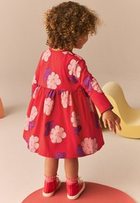 Rochie roșie cu mâneci lungi, cu modele mari de flori roz și mov, talie strânsă, cu o fustă evazată, asortată cu adidași roșii și șosete roz.