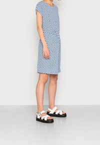 Robe rayée bleu et blanc à manches courtes, taille nouée ; associée à des sandales blanches avec semelles épaisses noires et lanières.