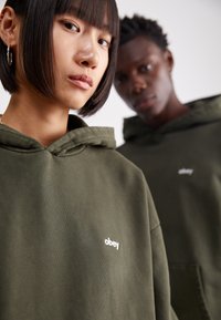 Sweatshirt à capuche vert olive avec une poche avant, arborant un petit logo "obey" blanc sur la poitrine. Tissu doux avec une coupe décontractée.
