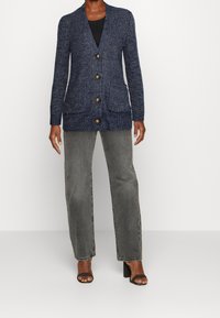 Cardigan lavorato a maglia blu navy con colletto a scialle, cinque bottoni marroni, due tasche frontali e polsini a coste, abbinato a jeans grigi scuri a gamba dritta.