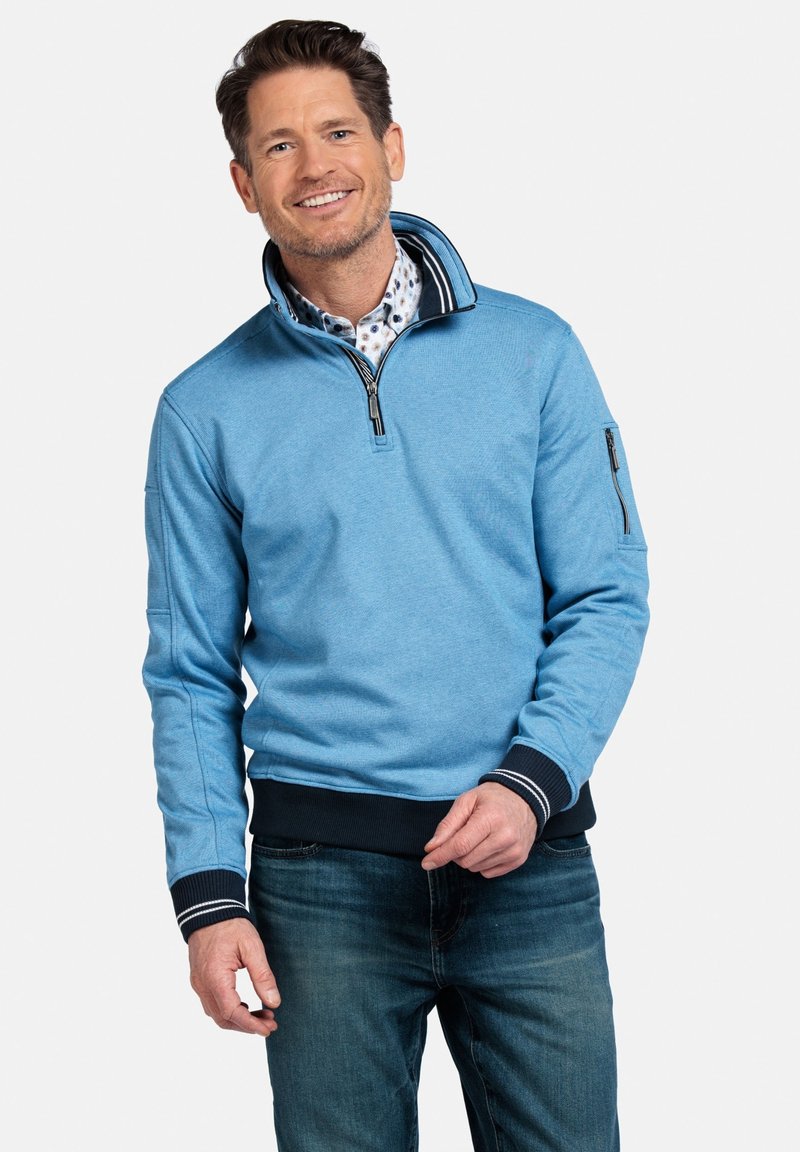 Lichtblauwe rits-hoodie met ribgebreide kraag, marineblauwe accenten en een ritszakje op de mouw, gedragen over een gestreept overhemd met kraag.