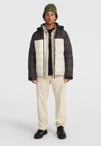 Pufferjack met zwarte en crèmekleurige secties, met capuchon en ritssluiting. Gelaagd beige shirt, cargobroek en zwarte wandelschoenen.