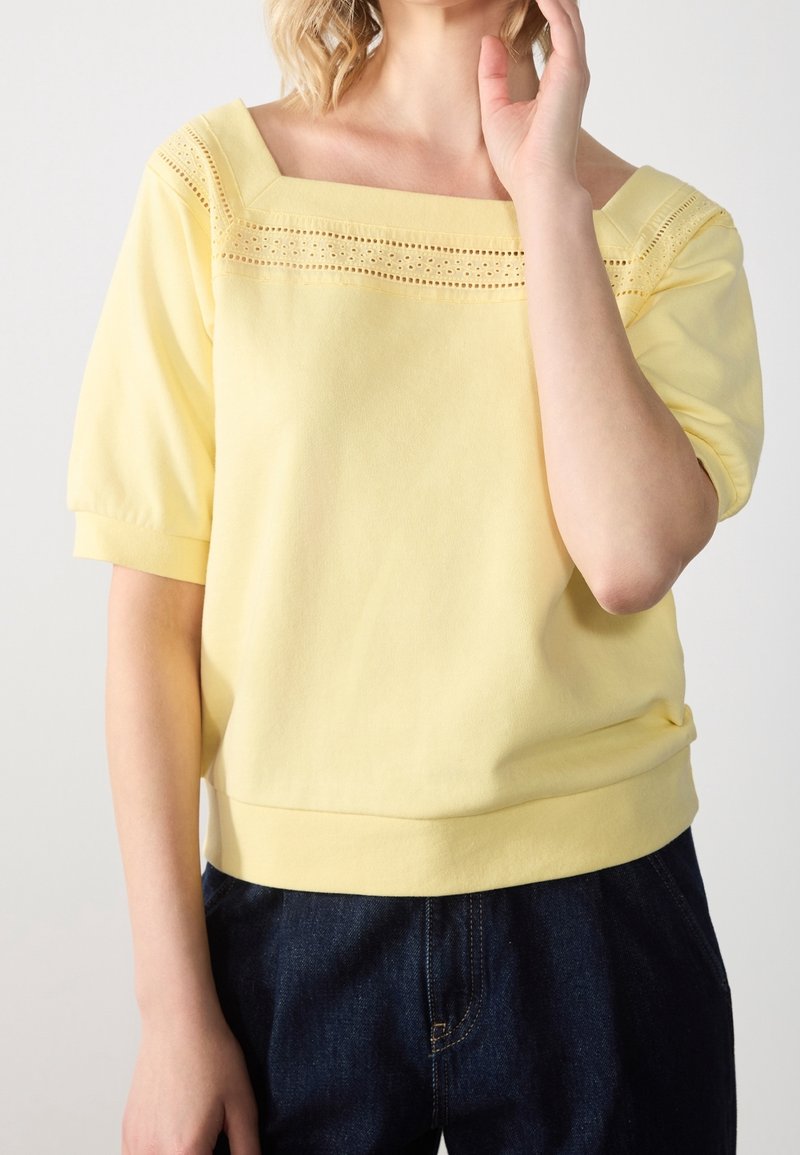 Femme portant un haut jaune pâle à encolure carrée avec bordure perforée et manches courtes bouffantes, associé à un pantalon en denim foncé.