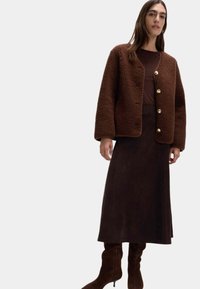 Giacca in pile marrone, testurizzata, con bottoni dorati, indossata sopra un top marrone scuro e una lunga gonna, abbinata a stivali marroni al ginocchio.