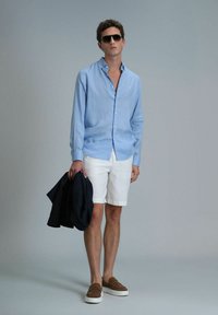 Hellblaues Leinenhemd, weiße Shorts, marineblaue Jacke in der Hand, braune Slipper, Sonnenbrille, Modell steht vor einem schlichten grauen Hintergrund.