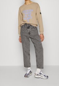 Sudadera beige con texto azul "PORQUE NO HAY PLANETA B", combinada con vaqueros grises de talle alto y zapatillas multicolores.