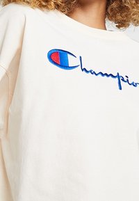 Cremefarbener Hoodie mit einem blauen und roten gestickten Logo. Lockerer Schnitt mit Rundhalsausschnitt und weichem Baumwollstoff.
