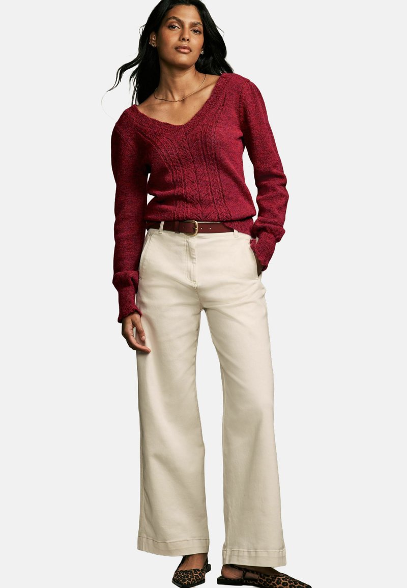 Maglione rosso a treccia con scollo a V, abbinato a pantaloni beige a gamba larga e scarpe nere con stampa leopardata, dotato di una cintura in pelle marrone.