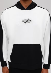 Sweat à capuche blanc avec des accents noirs, doté d'une poche avant et d'un logo circulaire sur la poitrine. Texture douce et coupe décontractée.