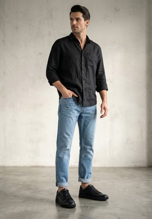 Mann trägt ein schwarzes Hemd mit Knöpfen, hellblaue hochgekrempelte Jeans und schwarze Sneaker, steht mit einer Hand in der Tasche und schaut zur Seite.