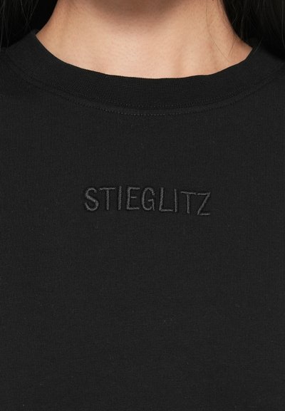 Fekete pamut póló bordás kerek nyakkal és hímzett "STIEGLITZ" logóval, amely egyszínű a szövet textúrájával.