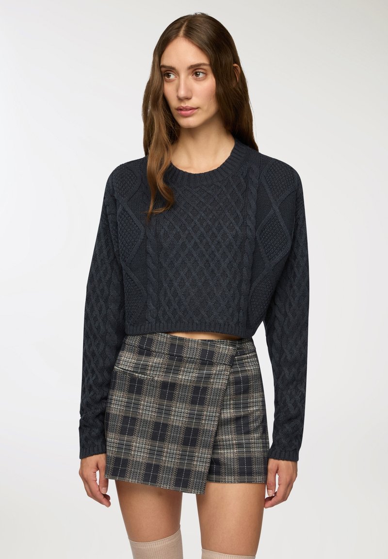 Maglione corto color navy con texture a treccia, abbinato a una mini gonna a quadretti. Il maglione presenta maniche lunghe e orlo a coste. Design classico.